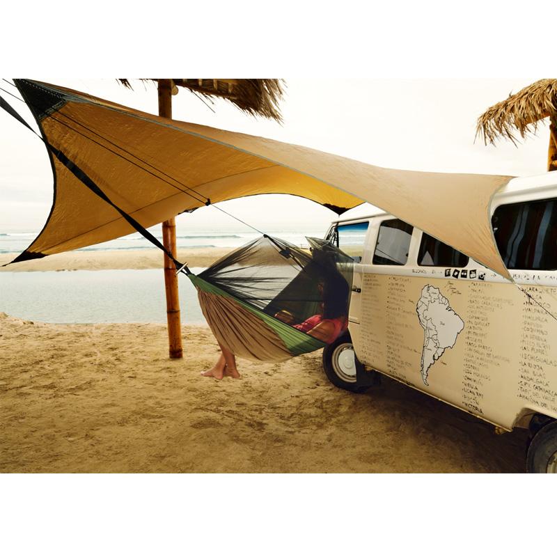 A lightweight Amazonas Adventure Ultra-Light Tarp is strung between a van and a palm tree on a sandy beach, providing shade. The van's side features a map of South America.