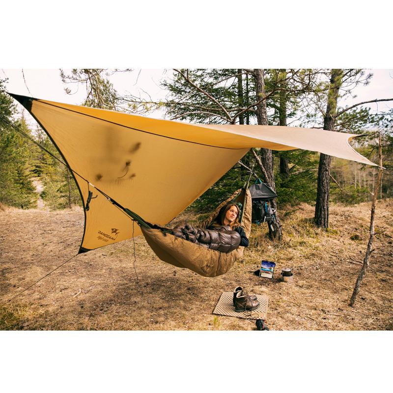 A person relaxes in an ultra-light hammock under a large yellow Amazonas Adventure Ultra-Light Tarp in a wooded area. Nearby, outdoor gear such as a backpack and hiking boots are visible. The scene is surrounded by trees with both grassy and forested terrain, all enveloped by the durable nylon ripstop fabric.