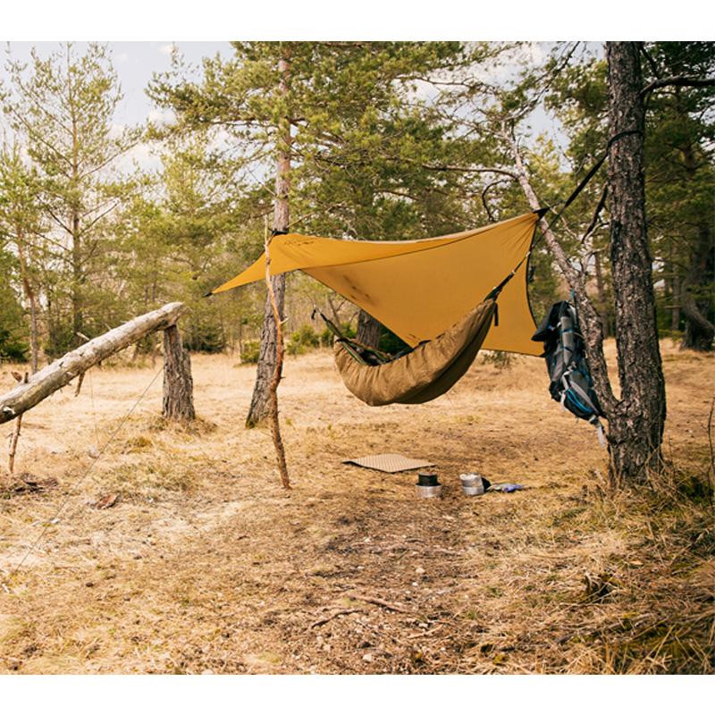 An ultra-light hammock and a sleeping bag hang elegantly between two trees in a grassy, wooded area. Overhead, an Amazonas Adventure Ultra-Light Tarp provides shelter, while nearby camping gear, including a backpack and pots, is arranged on the ground.