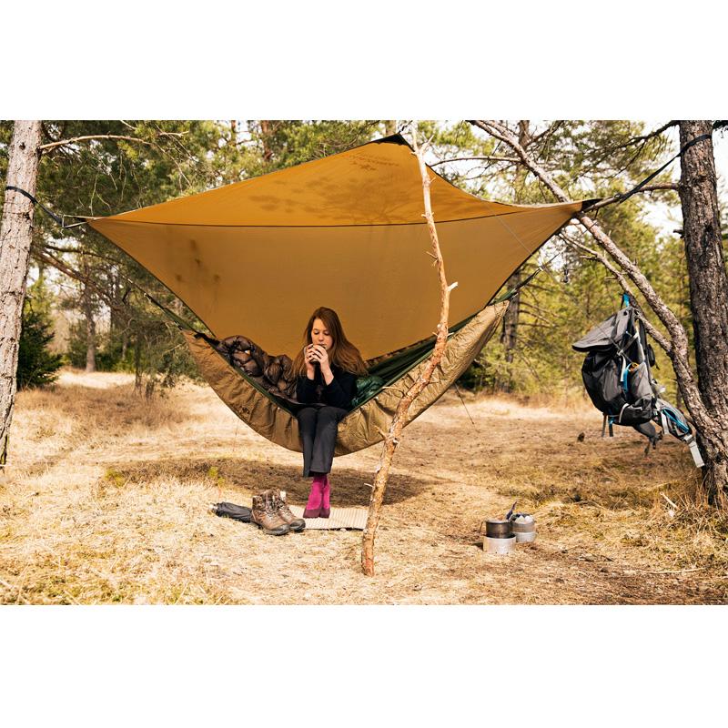 In the forest, someone relaxes in an ultra-light hammock beneath the Amazonas Adventure Ultra-Light Tarp, sipping from a mug. Nearby, their backpack and hiking gear hang from a tree. The ground is covered with dry grass, and the trees offer calming shade for this peaceful wilderness getaway.