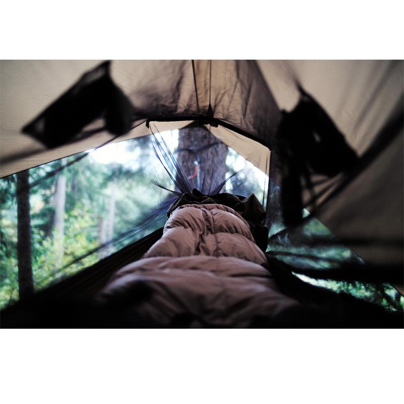 Inside the tent, a person lies comfortably in a sleeping bag, with the forest’s serene expanse visible just beyond the entrance. From the perspective at the foot of the sleeping bag, an Adventure Ultra-Light Tarp by Amazonas and durable Nylon Ripstop gear promise endless exploration opportunities.