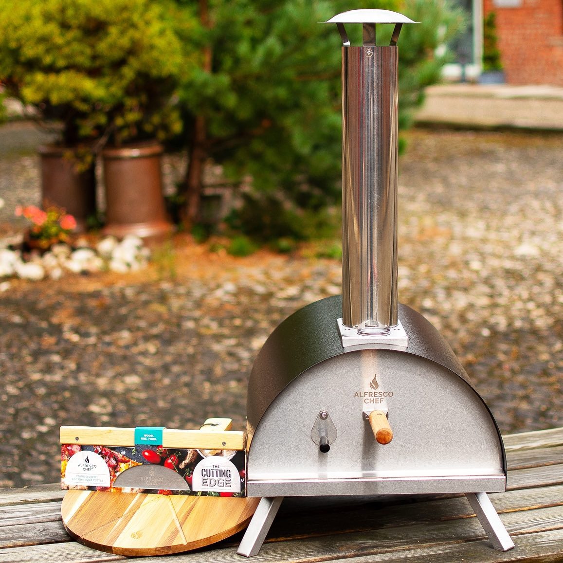 The Alfresco Chef Commis Chef Bundle, a sleek outdoor pizza oven with a metal chimney, stands on a stone patio. A wooden board with Cutting Edge Firewood sits to the left. Green bushes and trees provide a vibrant backdrop, making it perfect for an alfresco cooking experience.