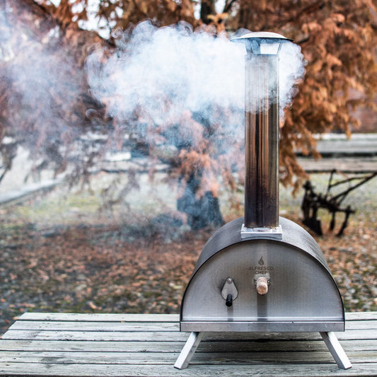 A compact metal Alfresco Chef Commis Bundle Oven rests on a wooden table, its chimney gently emitting smoke. This portable oven uses wood pellets to craft perfect pizzas, set against a backdrop of autumn trees with rich brown leaves.