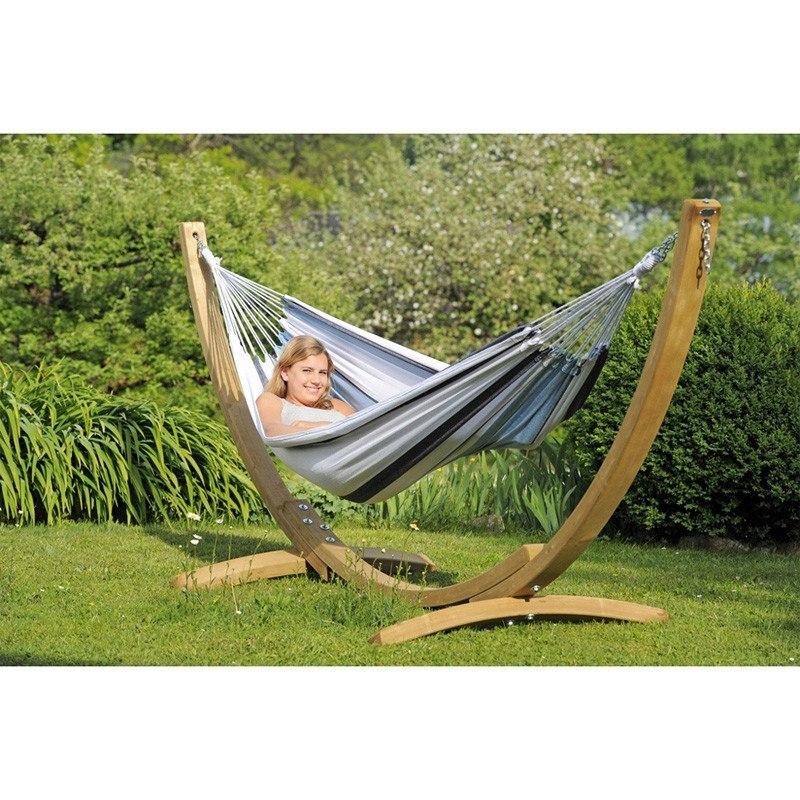 A person lounging in the Apollo & Marine Hammock Set by Amazonas, crafted from sustainable European Spruce and supported by a wooden stand, creates a serene atmosphere in a lush garden. Surrounded by vibrant shrubs and trees, this inviting hammock set becomes the centerpiece of tranquility.