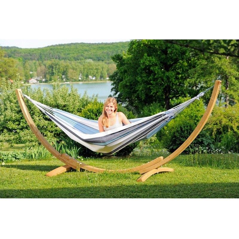 A person smiles while lounging in the Apollo & Marine Hammock Set by Amazonas, crafted from sustainable European Spruce, on a grassy lawn. In the background, a scenic view of trees, a lake, and distant greenery under a clear sky enhances the setting—truly making this Amazonas hammock a garden centerpiece.