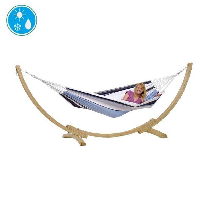 A woman smiles while relaxing in the Apollo & Marine Hammock Set by Amazonas, featuring a blue and white striped hammock crafted from sustainable European Spruce, elegantly suspended on a wooden stand. Icons in the top left emphasize its all-weather suitability, making it an ideal focal point for any garden.