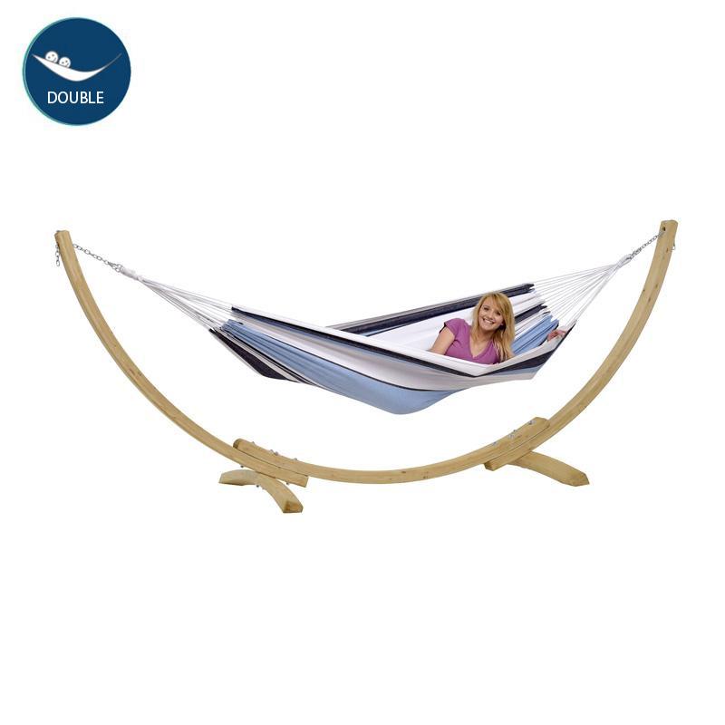 A woman is lying in the Amazonas Apollo & Marine Hammock Set, a striking garden centerpiece. She is smiling and appears relaxed in the double hammock with a wooden stand. The hammock's design features white, blue, and gray stripes crafted from sustainable European Spruce. A DOUBLE label can be seen in the corner.