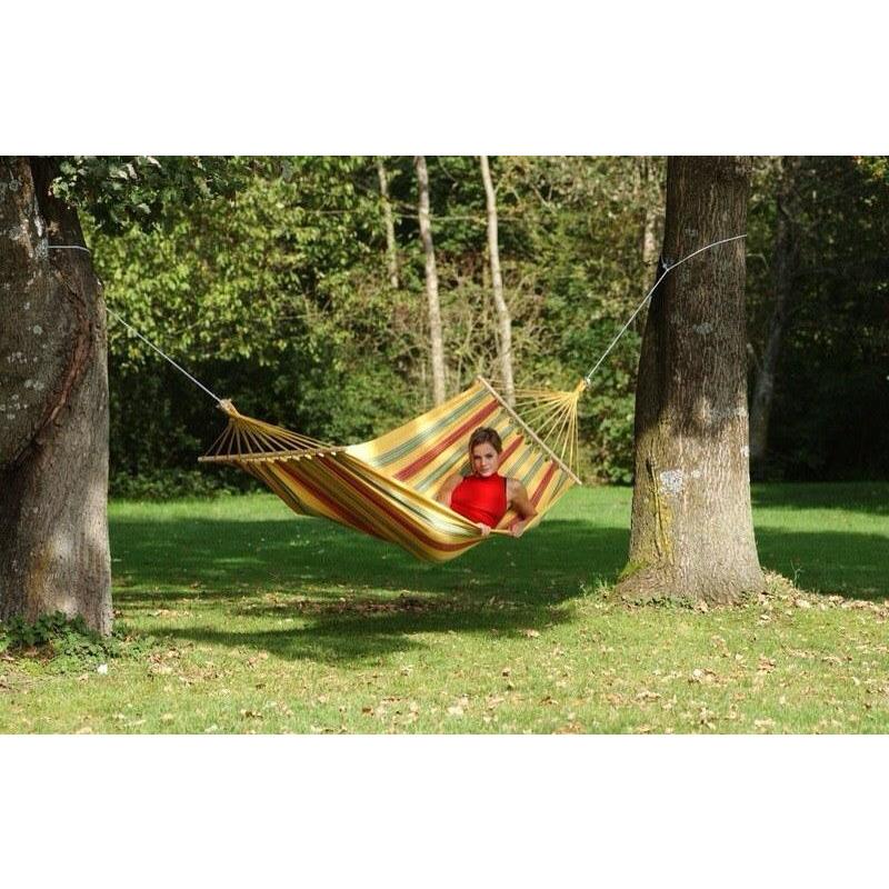 A person is unwinding in the Amazonas Aruba Vanilla Hammock, securely tied between two trees in a grassy area adorned with scattered leaves. The background displays vibrant greenery under a bright, clear sky, highlighting an ideal combination of relaxation and style through this garden furniture hammock setup.