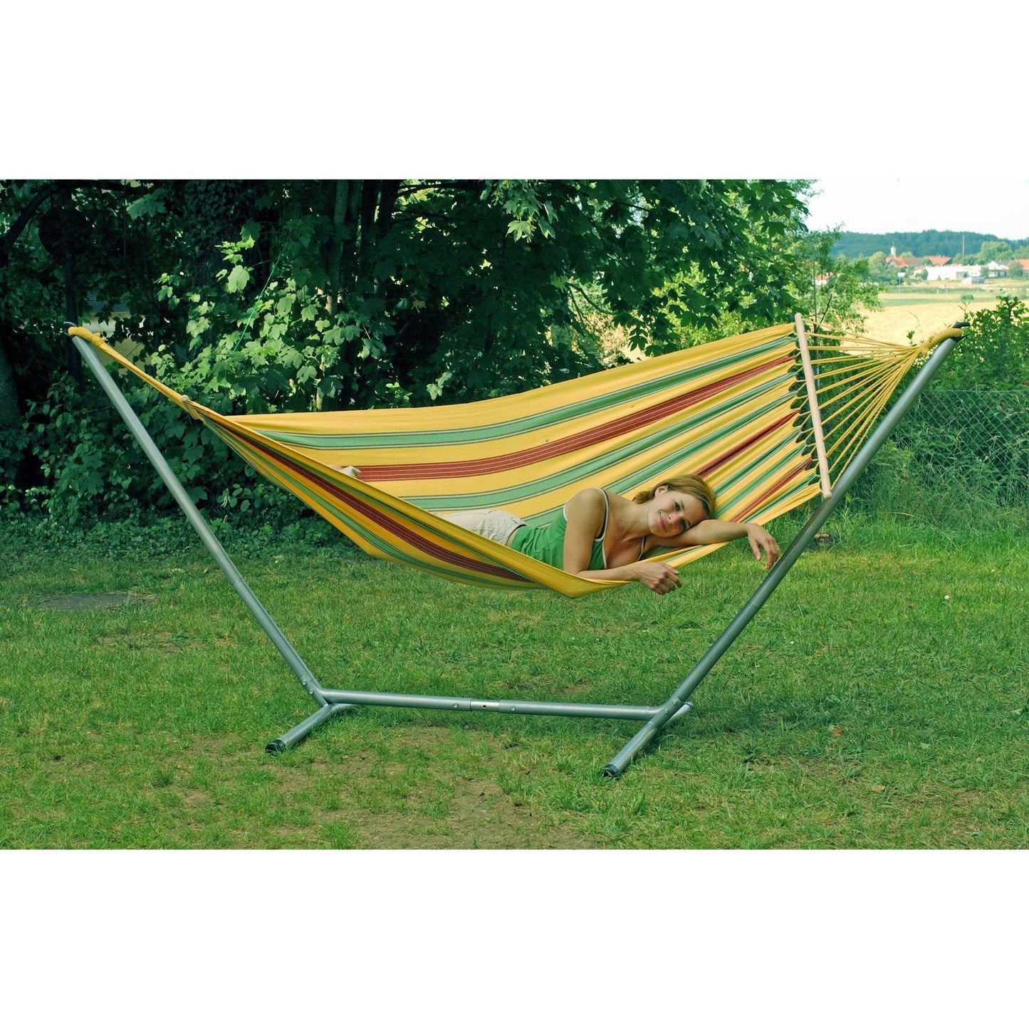 A person relaxes on an Amazonas Aruba Vanilla Hammock in a grassy backyard. The weatherproof hammock is supported by a metal stand, with lush green trees in the background, creating a peaceful outdoor setting.