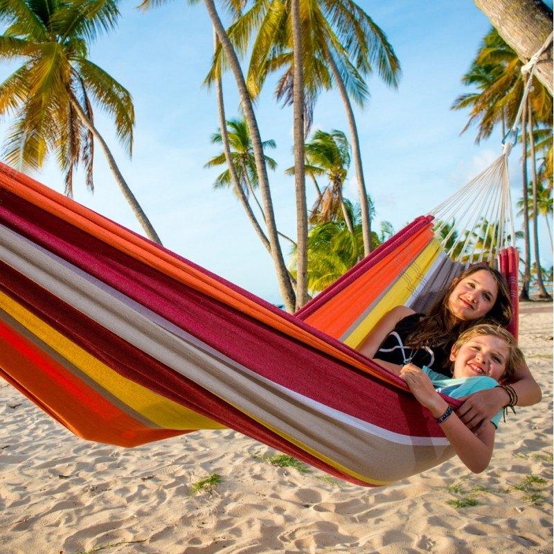 Two people smiling and relaxing in a colorful, durable Barbados Acerola Hammock by Amazonas on a sandy beach, surrounded by tall palm trees under a clear blue sky.