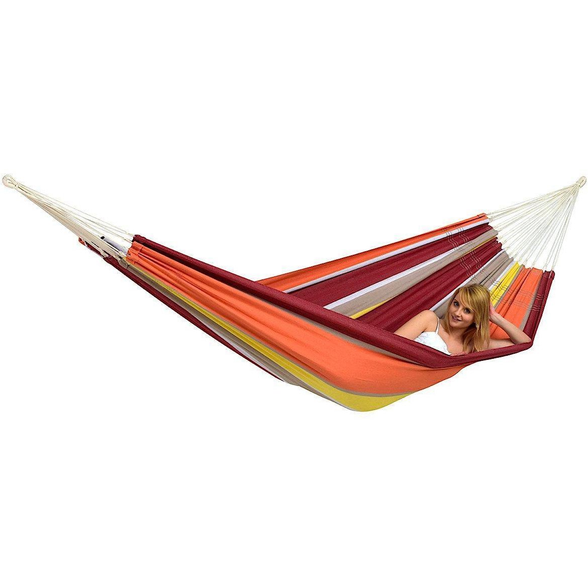 A person lounging in the Barbados Acerola Hammock by Amazonas, crafted from durable Brazilian cotton and showcasing vibrant red, orange, yellow, and white stripes against a white background. The hammock is securely suspended at both ends, allowing the person to smile and recline comfortably.