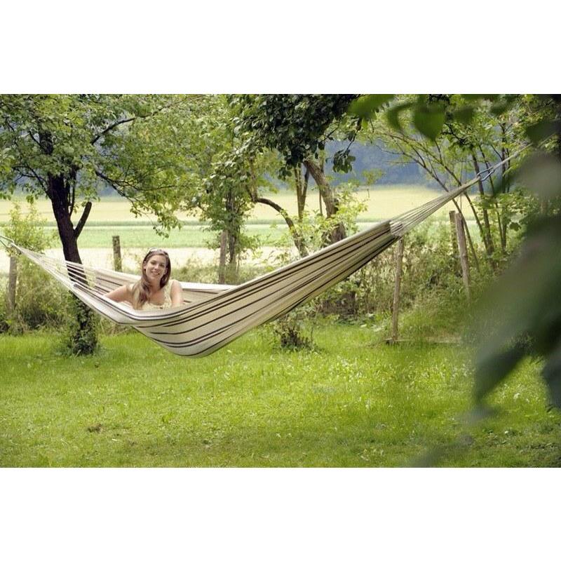 A person unwinds in the spacious Barbados Cappuccino Hammock by Amazonas, crafted from premium Brazilian cotton, gently swaying between trees in a vibrant, green garden. The wooden fence and open field in the background enhance the sun-drenched scene's peacefulness and serenity.