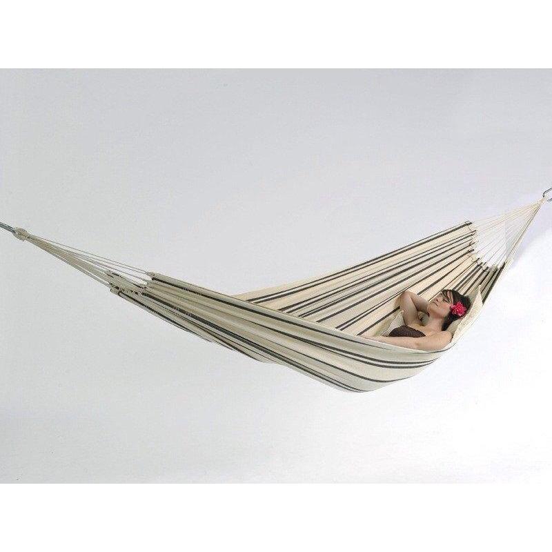 A person is relaxing in a generously sized hammock, resting one arm behind their head. Hanging in a simple white setting, the Barbados Cappuccino Hammock from Amazonas pairs perfectly with their outfit and sunglasses as a flower decorates their hair.