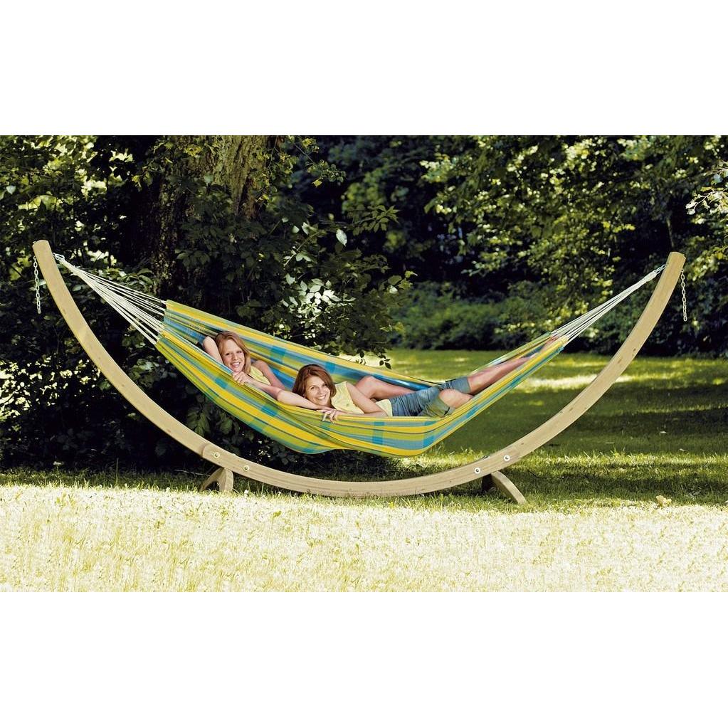 Two individuals unwind in a colorful Amazonas Barbados Lemon Hammock crafted from Brazilian cotton, suspended by a wooden frame in a sunlit garden. They rest side by side, smiling amid lush green trees and grass.