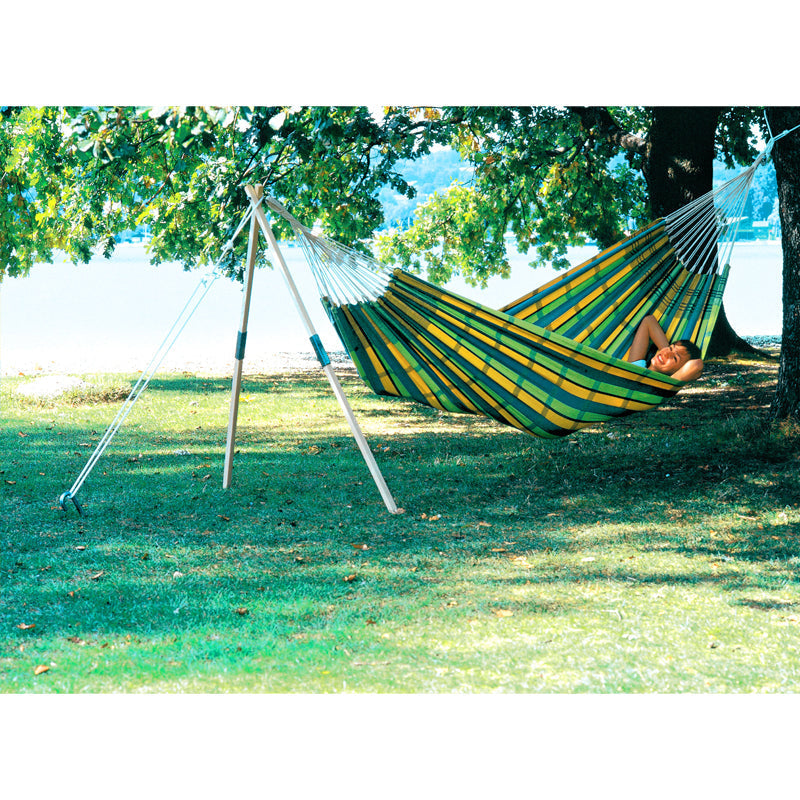 A person unwinds in an Amazonas Barbados Lemon Hammock under the shade of a large tree. The lush green grass complements the peaceful lakeside view in the background.