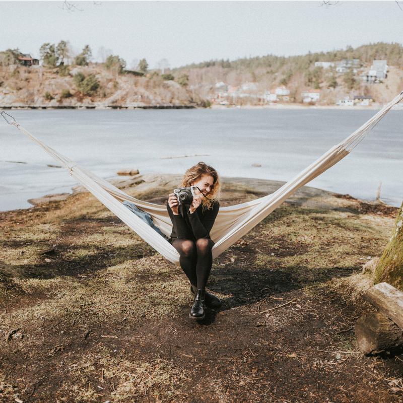 A person sits in an Amazonas Barbados Natura Hammock by a lake, holding a vintage camera. The landscape features a calm body of water, gentle hills, and trees in the background. Smiling and relaxed in this natural setting, they embrace the serenity around them.