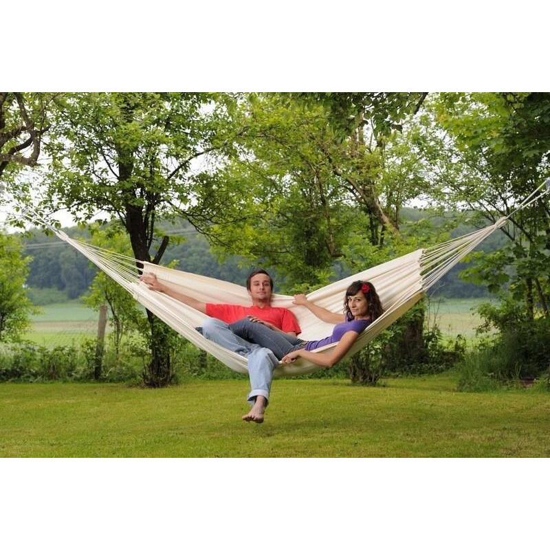 A couple relaxes in an Amazonas Barbados Natura Hammock, crafted from durable Brazilian cotton and suspended between two trees in a lush garden. The setting features green grass and leafy trees, with a backdrop of more trees and open fields, creating the perfect family moment in this serene environment.