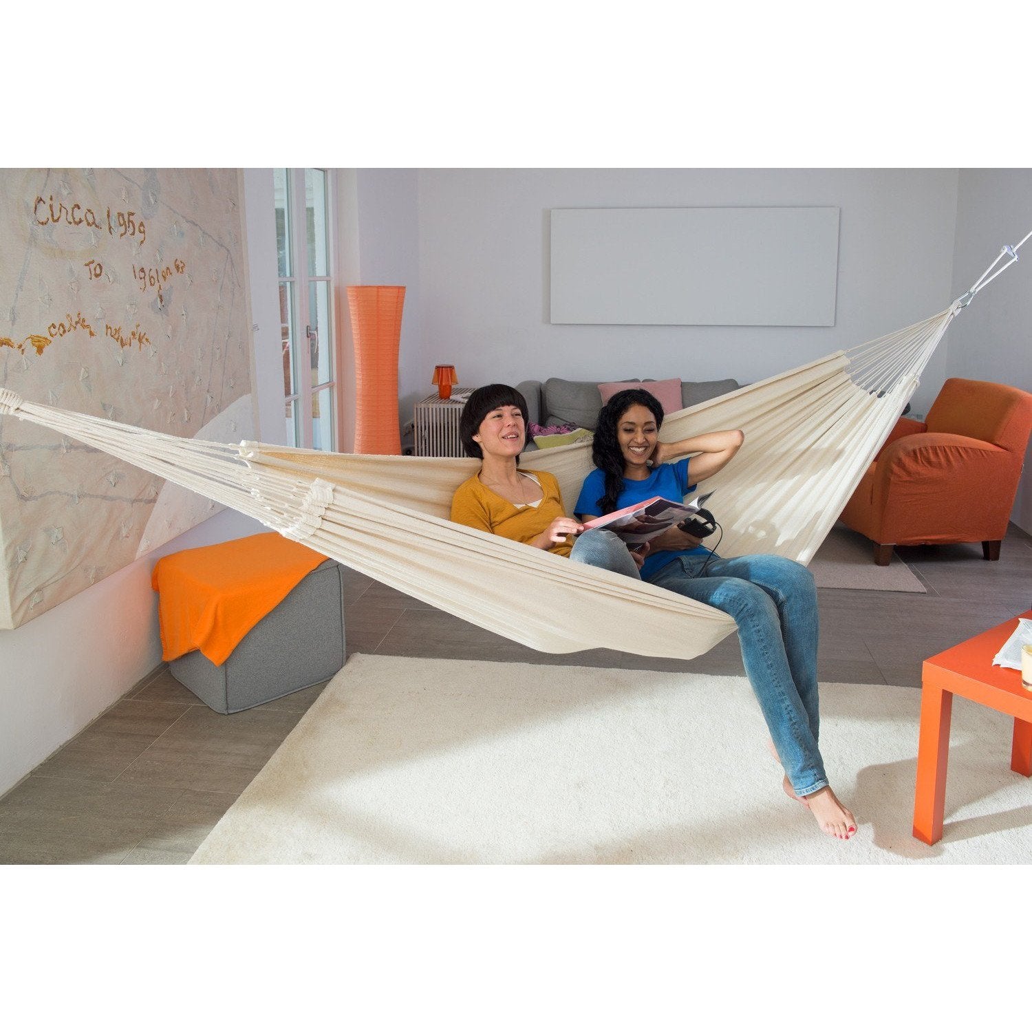 Two people are enjoying a relaxing moment in an Amazonas Barbados Natura Hammock inside. One person is engrossed in a book, and both are smiling, surrounded by comfort. The cozy room includes a couch, an orange chair, and striking wall art, all illuminated by bright daylight.