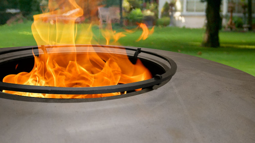 A close-up view of the BonFeu Foodbumper by Bonfeu, showcasing its sleek steel ring with bright orange flames dancing around it. In the background, a lush green lawn, towering trees, and a cozy house complete the picturesque scene.