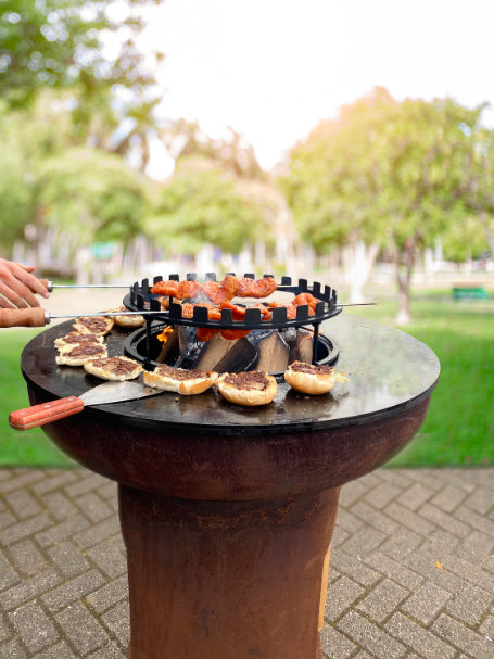 A round Bonfeu Skewers Ring sizzles with bagels and skewered meat cooking on its steel surface. Two hands carefully adjust the skewers, while the blurred greenery and trees in the park setting create a serene backdrop.