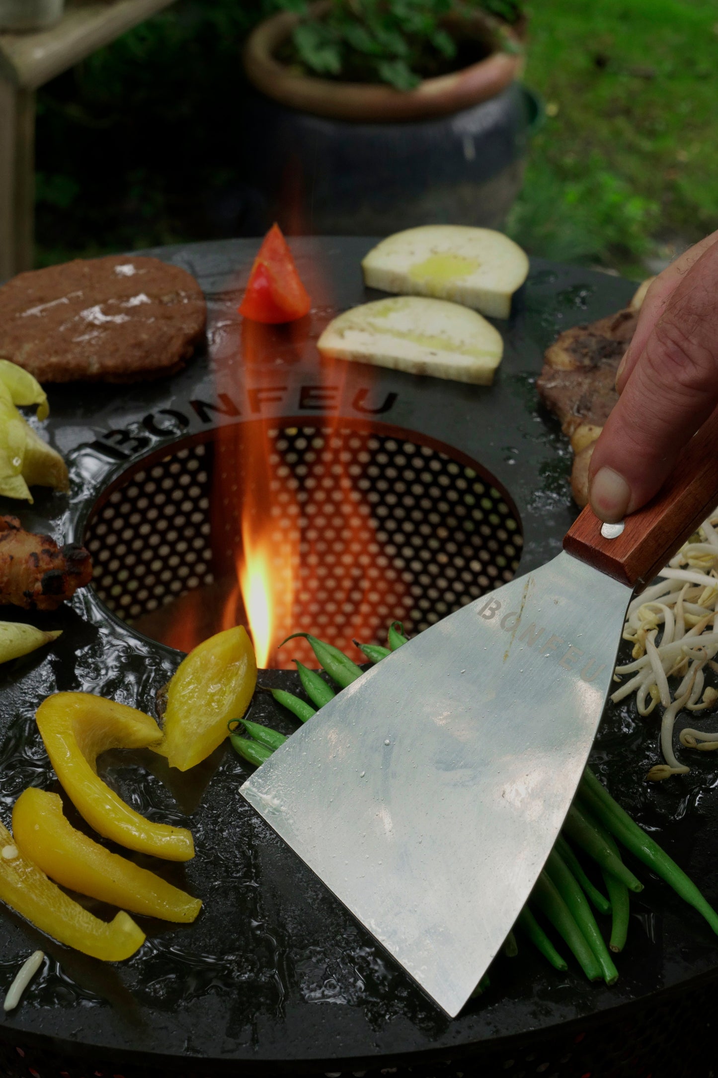 A hand holds a Bonfeu BonFeu Spatula with a wooden handle over a sizzling grill filled with yellow peppers, green beans, potatoes, and a slice of tomato. Nearby, a burger patty and sprouts are also cooking. The stainless-steel blade shines in the outdoor setting as a small flame flickers in the center.