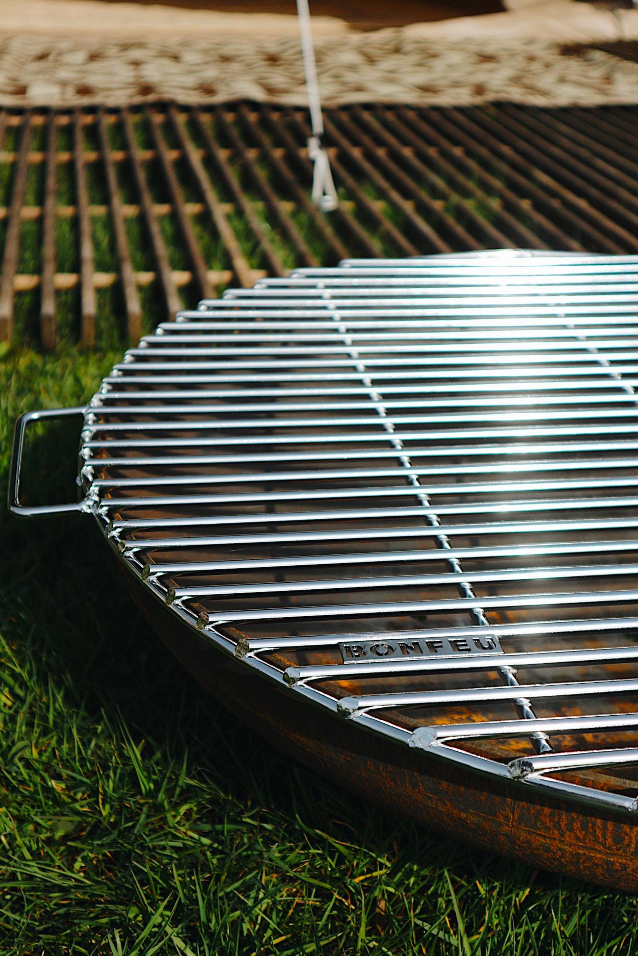 The sentence can be rewritten as: A close-up of a shiny, steel Bongrill full circle by Bonfeu sits on a grassy surface. The word BONFEU is engraved on the edge of the grill. Wooden planks are visible in the background, slightly blurred.