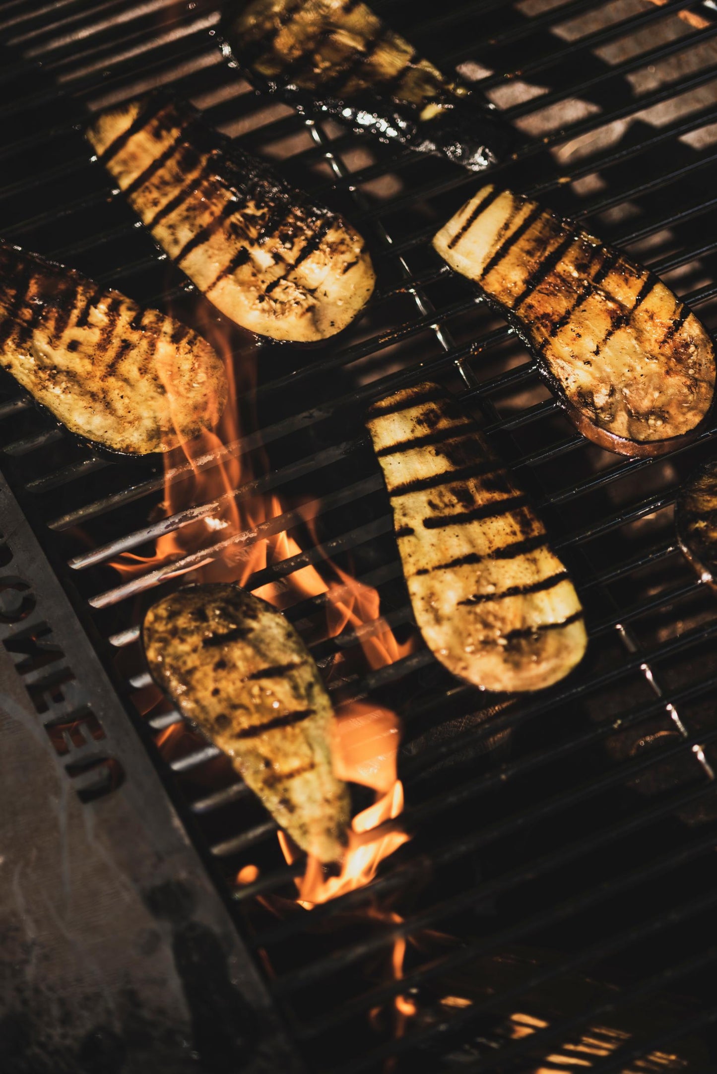 Grilled slices of eggplant with char marks on a Bonfeu Bongrill Full Circle barbecue over open flames create a rustic and appetizing appearance. The eggplant, seasoned to perfection, shows a golden-brown finish against the sleek steel grill.