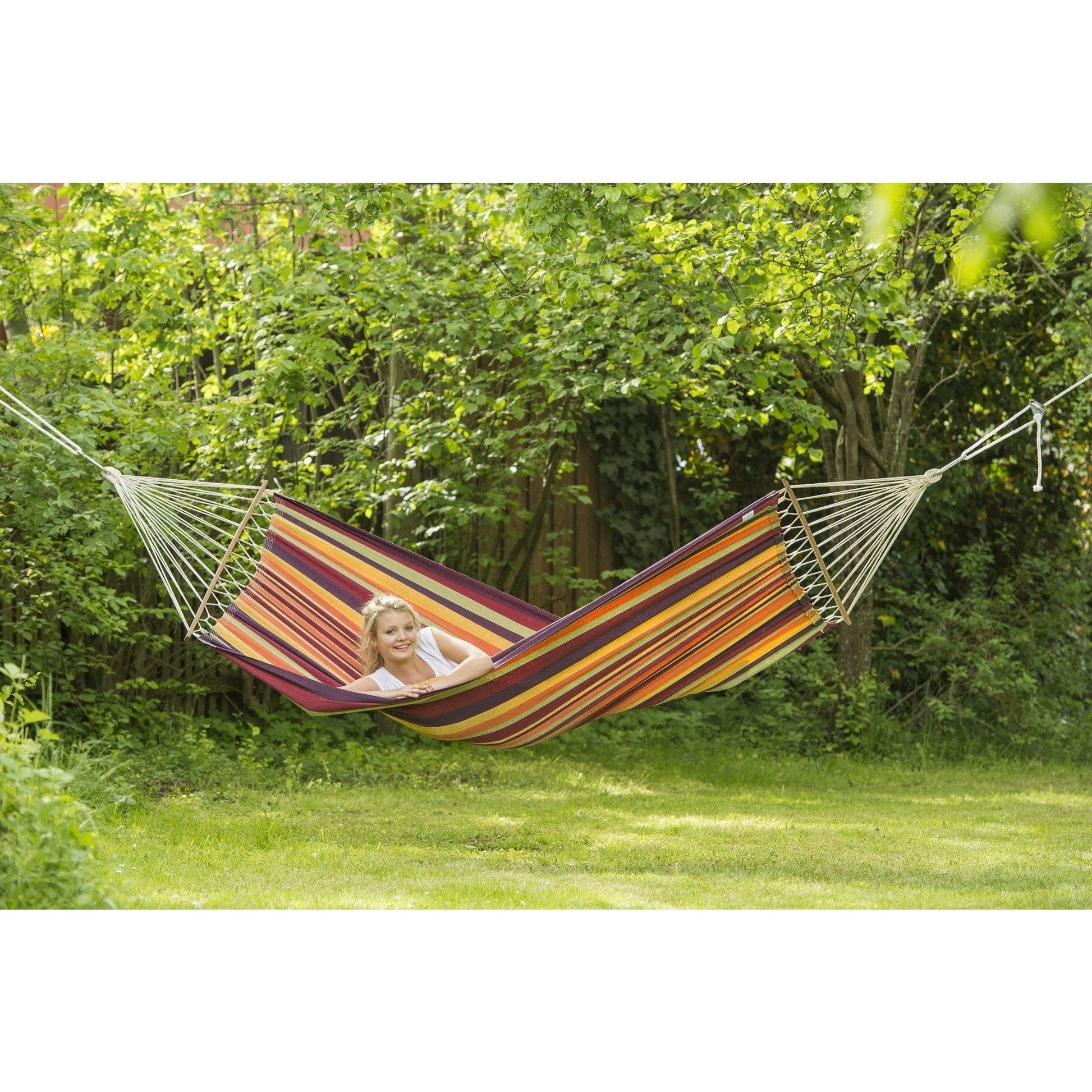 In a lush, green garden, a person relaxes in the vibrant Brasilia Tropical Hammock by Amazonas, made from soft Brazilian cotton and suspended between two trees. Sunlight filters through the leaves, crafting a tranquil outdoor atmosphere.