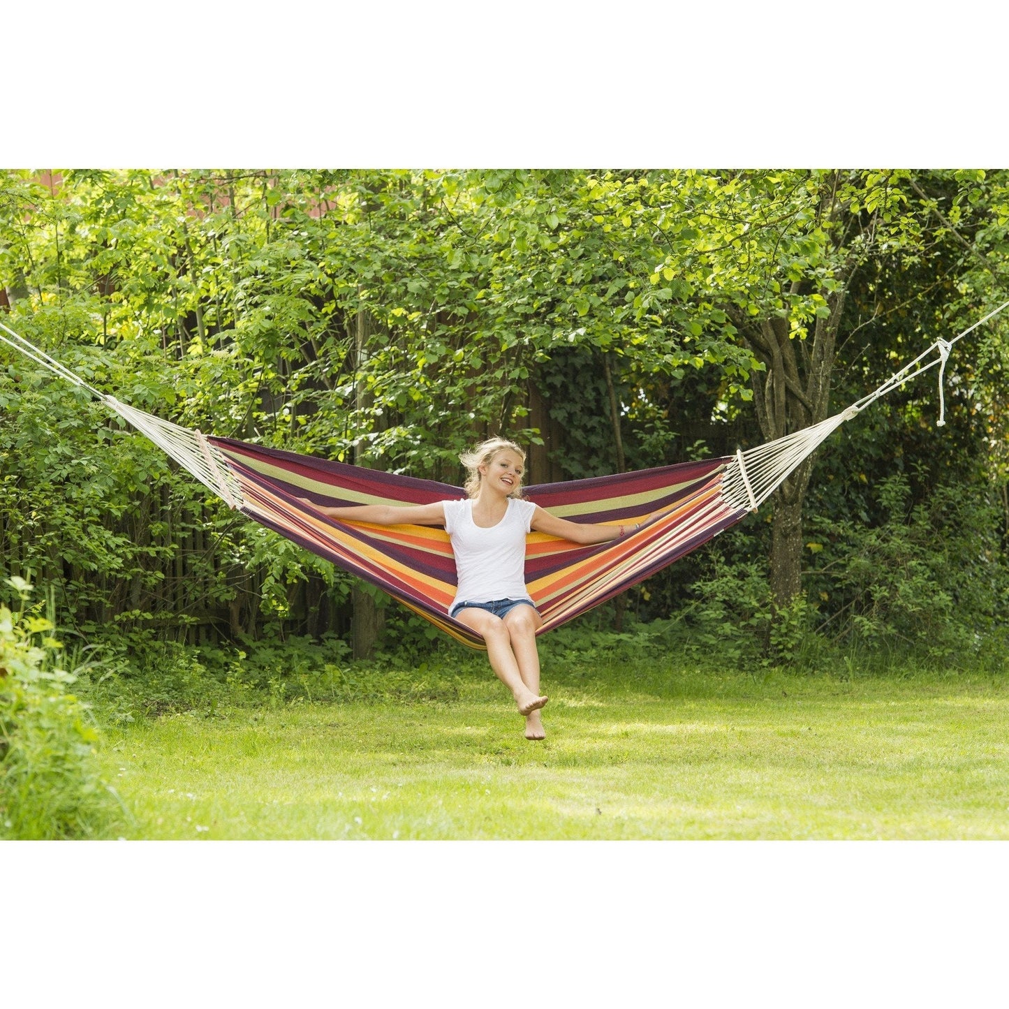 A person in a white t-shirt and shorts sits comfortably in the Amazonas Brasilia Tropical Hammock, smiling. The vibrant hammock is suspended between trees, surrounded by lush green grass and foliage, creating a serene outdoor setting.