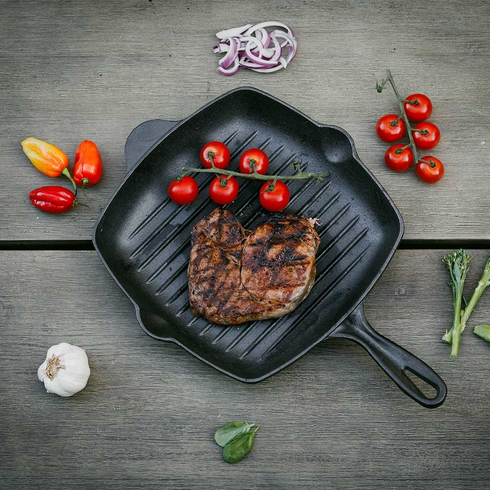 A heart-shaped grilled steak sizzles on the Casa Mia Cast Iron Skillet, celebrated for its excellent heat retention. It's accompanied by vibrant cherry tomatoes, colorful bell peppers, garlic, broccoli, spinach leaves, and sliced red onion on a wooden surface.