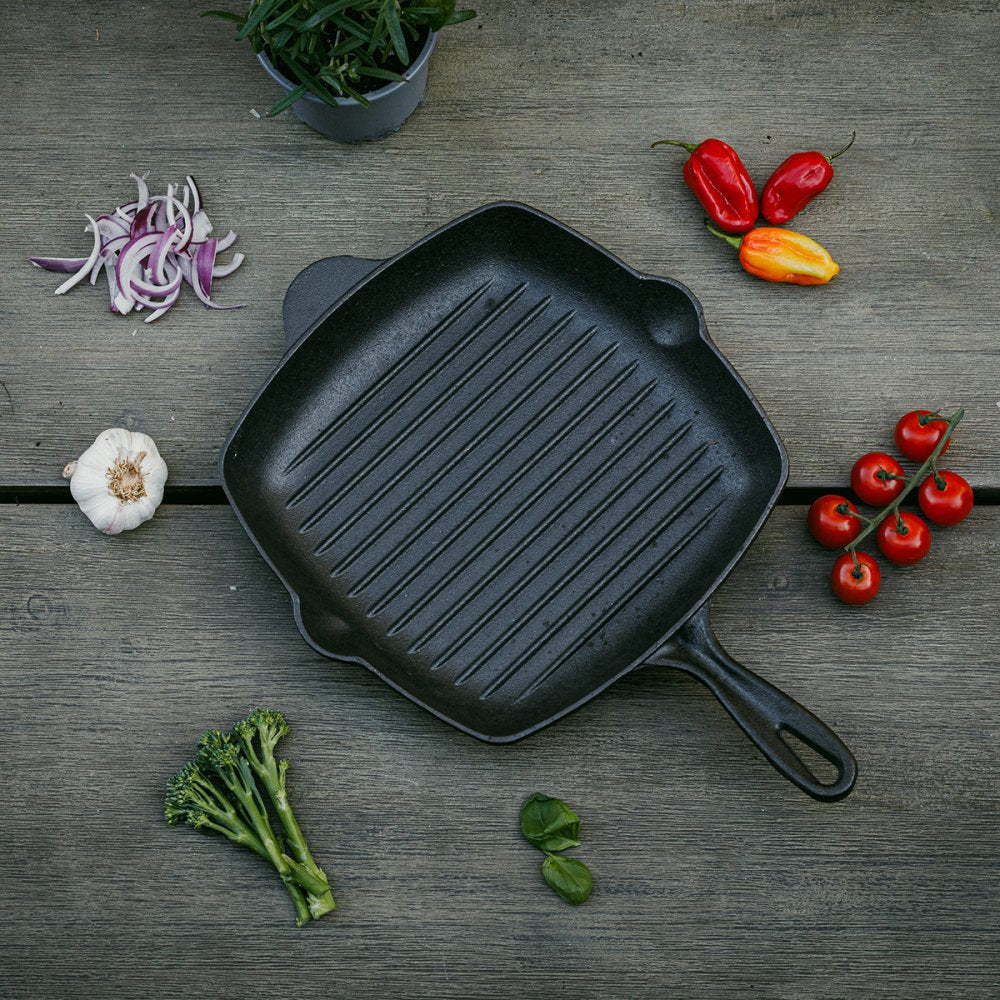 The Casa Mia Cast Iron Skillet sits on a wooden surface, encircled by vivid red onions, whole garlic cloves, crisp green broccoli, fresh basil leaves, and an assortment of red and yellow peppers. Nearby, cherry tomatoes remain on the vine with a pot of lush green foliage providing a rich backdrop.