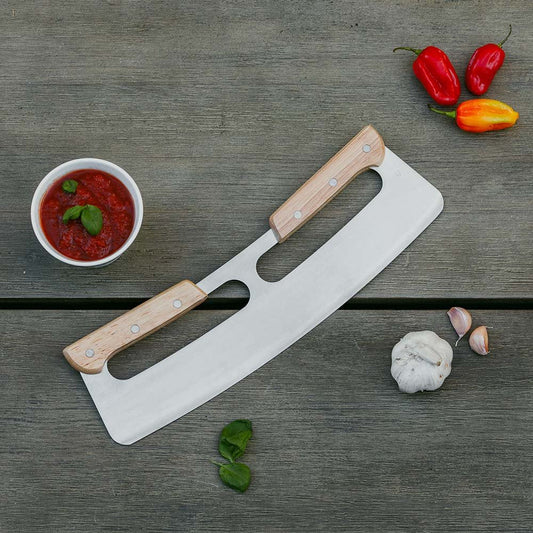 The Casa Mia Large Pizza Cutter, featuring a curved design and wooden handles, rests on a wooden surface, highlighting its ergonomic craftsmanship. Nearby are three small red and yellow peppers, a clove of garlic, fresh basil leaves, and a bowl of red sauce.