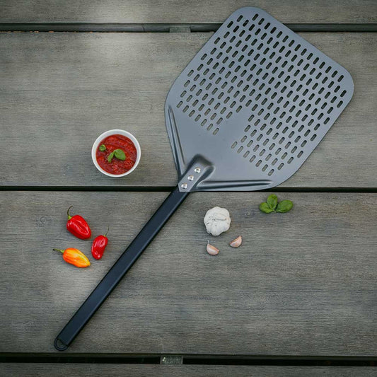 A Casa Mia Perforated Peel, featuring a sleek ergonomic design and a black handle, rests on a wooden surface. Nearby are red and yellow peppers, a bowl of tomato sauce, garlic cloves, and fresh basil leaves—perfect companions for creations from your favorite pizza ovens.