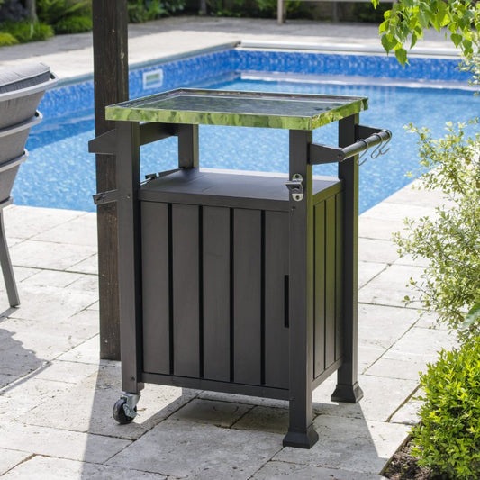 A Casa Mia Prep/Cooking Table with a black wood-paneled cabinet and stainless steel top is stationed by the patio near a pool. The table features plenty of storage space, hooks for utensils, and wheels for convenient mobility.