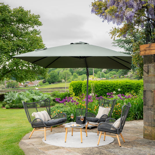 The outdoor seating area includes three wicker chairs surrounding a small round table with drinks, all shaded by the large Challenger T2 3m Square Olive Free Arm Parasol from Pacific Lifestyle. This peaceful garden setting is complemented by lush grass, blooming flowers, and trees in the background, adding to the tranquil atmosphere.