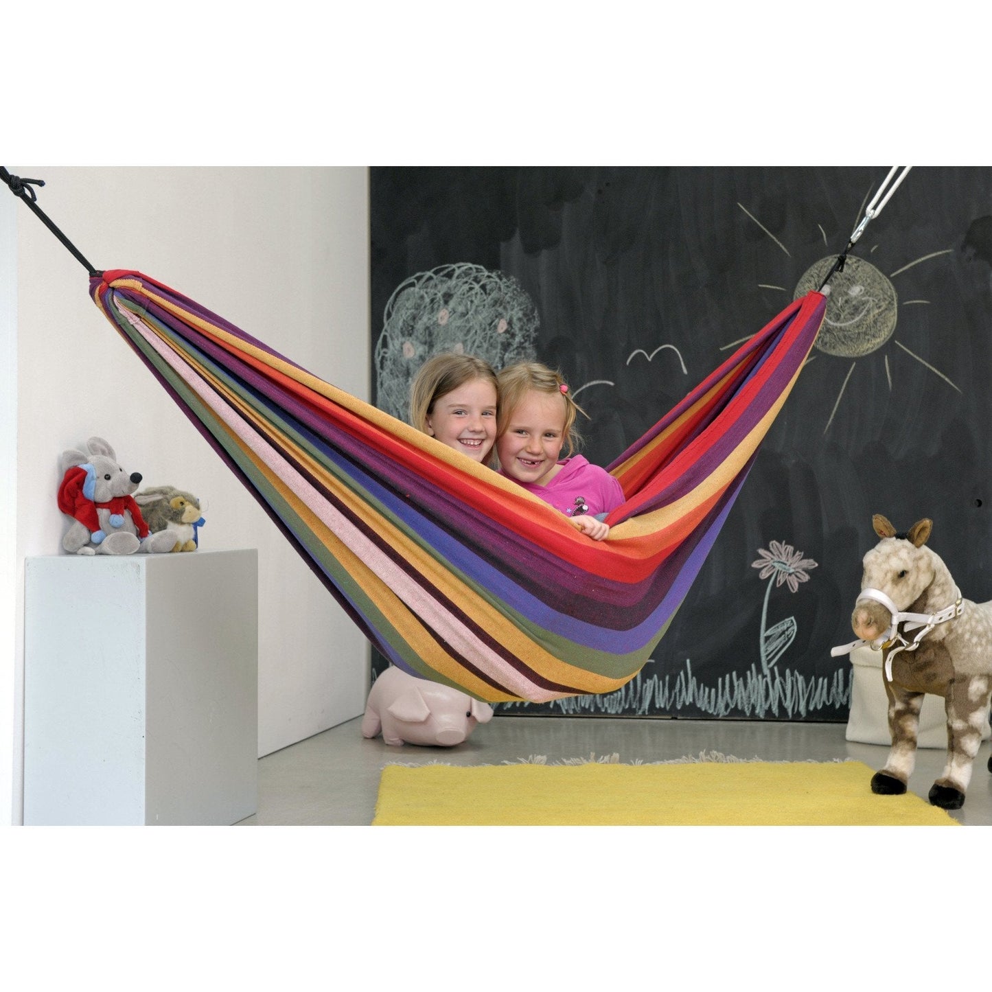 In a bright, vibrant setting, two young children are smiling as they sit in an Amazonas Chico Rainbow Hammock designed for children. The indoor background showcases a blackboard adorned with chalk drawings of the sun and clouds. Around the room, plush toys such as a horse and an elephant add to the playful atmosphere.