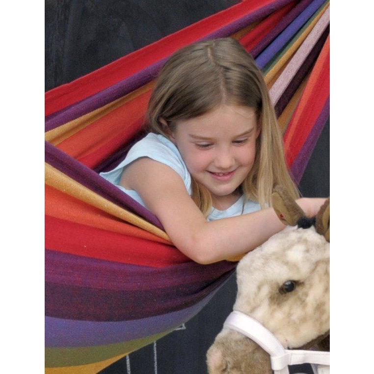 A young girl with long blonde hair smiles while lying in an Amazonas Chico Rainbow Hammock for children. She is gently petting a plush horse. The scene is cozy and cheerful, perfect for young children to enjoy their weatherproof outdoor adventures.