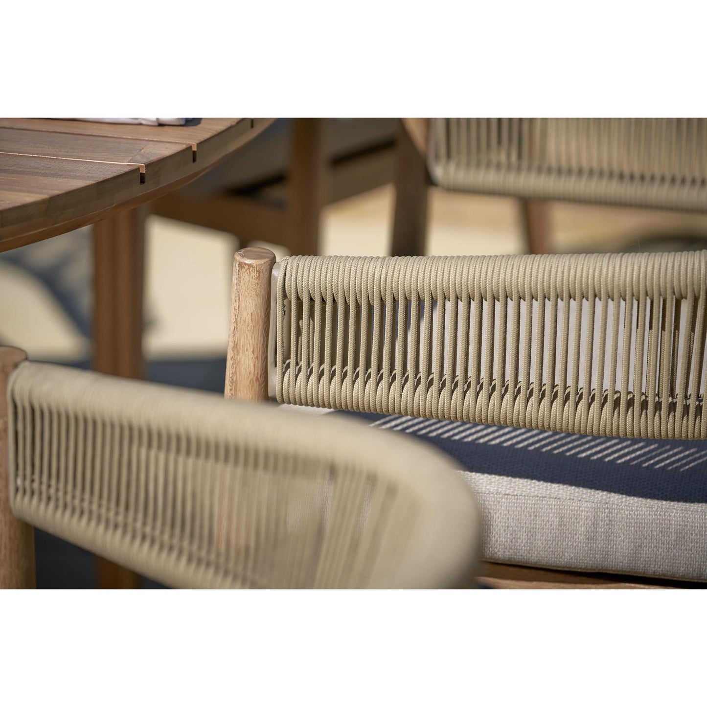 A close-up of the Denver Sage Green 6 Seater Dining Set by Pacific Lifestyle reveals wooden chairs with woven backrests and neutral cushions. Crafted from Acacia wood, this outdoor dining set includes a matching table, partially visible in the scene, creating a cozy setting. Handcrafted in Vietnam, it exudes an artisanal charm perfect for outdoor living.