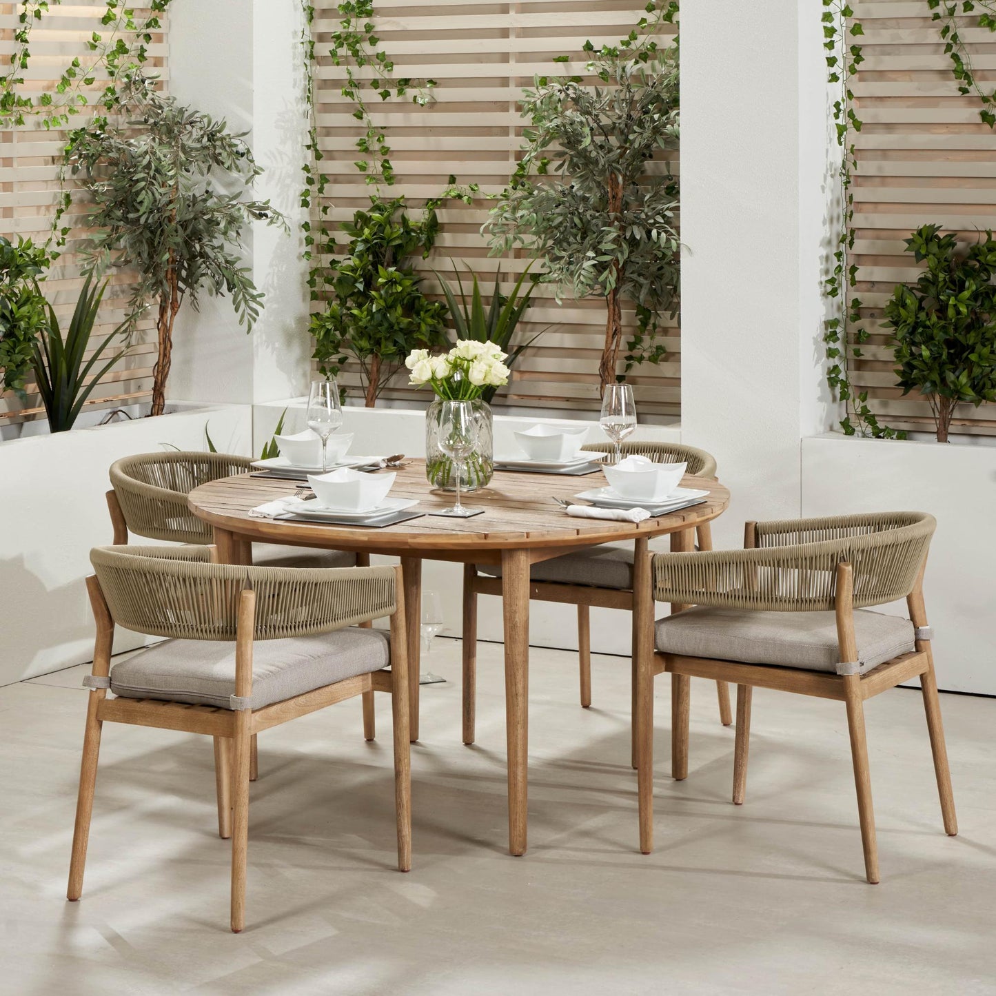 Introducing the Denver Sage Green 6 Seater Dining Set by Pacific Lifestyle, perfect for outdoor living and decor. This exquisite dining set is crafted in Vietnam and features a round acacia wood table complemented by six cushioned chairs. The table is beautifully styled with white dishes and a vase of white flowers, surrounded by lush green plants and wooden slatted panels.