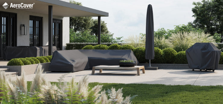 In a contemporary outdoor patio, grey furniture covers shield the sofas and a grill, all under the protection of an Aero Cover Free Arm Parasol Aerocover 240 x 68cm. Nearby stands a free arm parasol amidst thriving greenery and ornamental grasses. A wooden coffee table with a decorative bowl adds to the charm on the light-colored paved area.