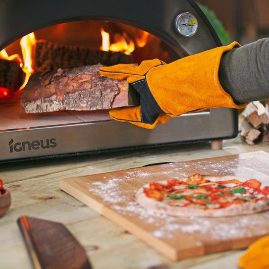 A person wearing Igneus Heat Resistant Gloves places a log into the pizza oven. Nearby, a wooden cutting board holds a prepared pizza topped with tomatoes and herbs, while the visible fire from the oven creates a warm ambiance.