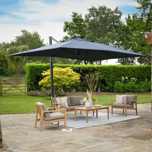 A spacious patio features a Pacific Lifestyle Icon Premium T1 4mx3m Rectangular Faded Black Parasol shading a seating area with a wooden outdoor sofa, armchairs, and a coffee table on a rug. The premium-class fabric of the parasol complements the setting surrounded by green hedges and trees under a partly cloudy sky.