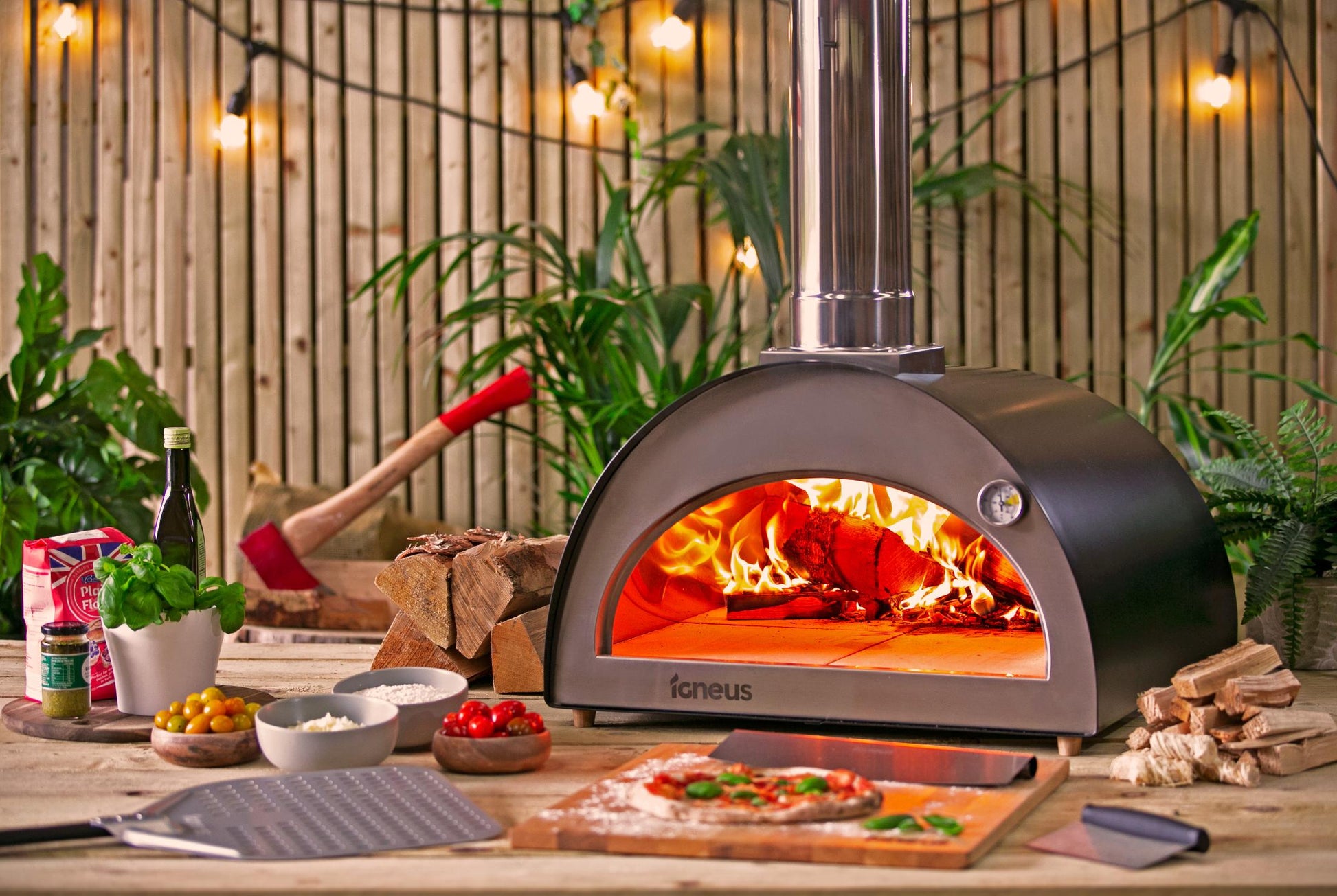 An Igneus Classico Pizza Oven Ultimate Bundle rests on a wooden table, its flames casting a cozy glow. Fresh ingredients such as tomatoes, herbs, and cheese are arranged around it, with dough ready for toppings. The enchanting scene is complemented by string lights and vibrant greenery, enhancing the outdoor cooking experience.