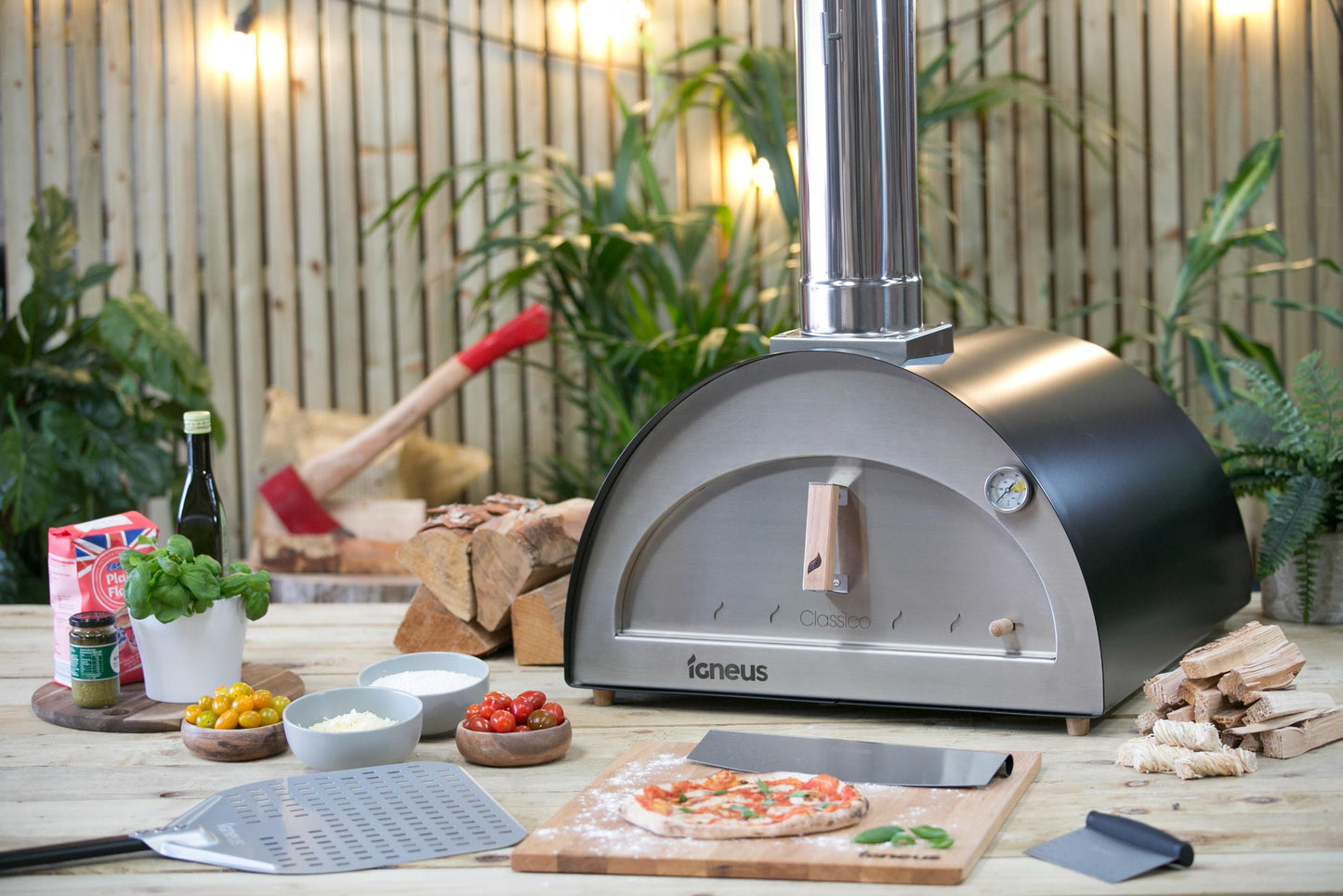 An Igneus Classico Pizza Oven Ultimate Bundle from the Igneus brand is placed on a wooden table, surrounded by ingredients such as tomatoes, cheese, basil, and olive oil. A pizza rests on a peel ready for baking. The scene is set against a backdrop of plants and warm lighting, creating an ideal atmosphere for outdoor cooking enthusiasts.