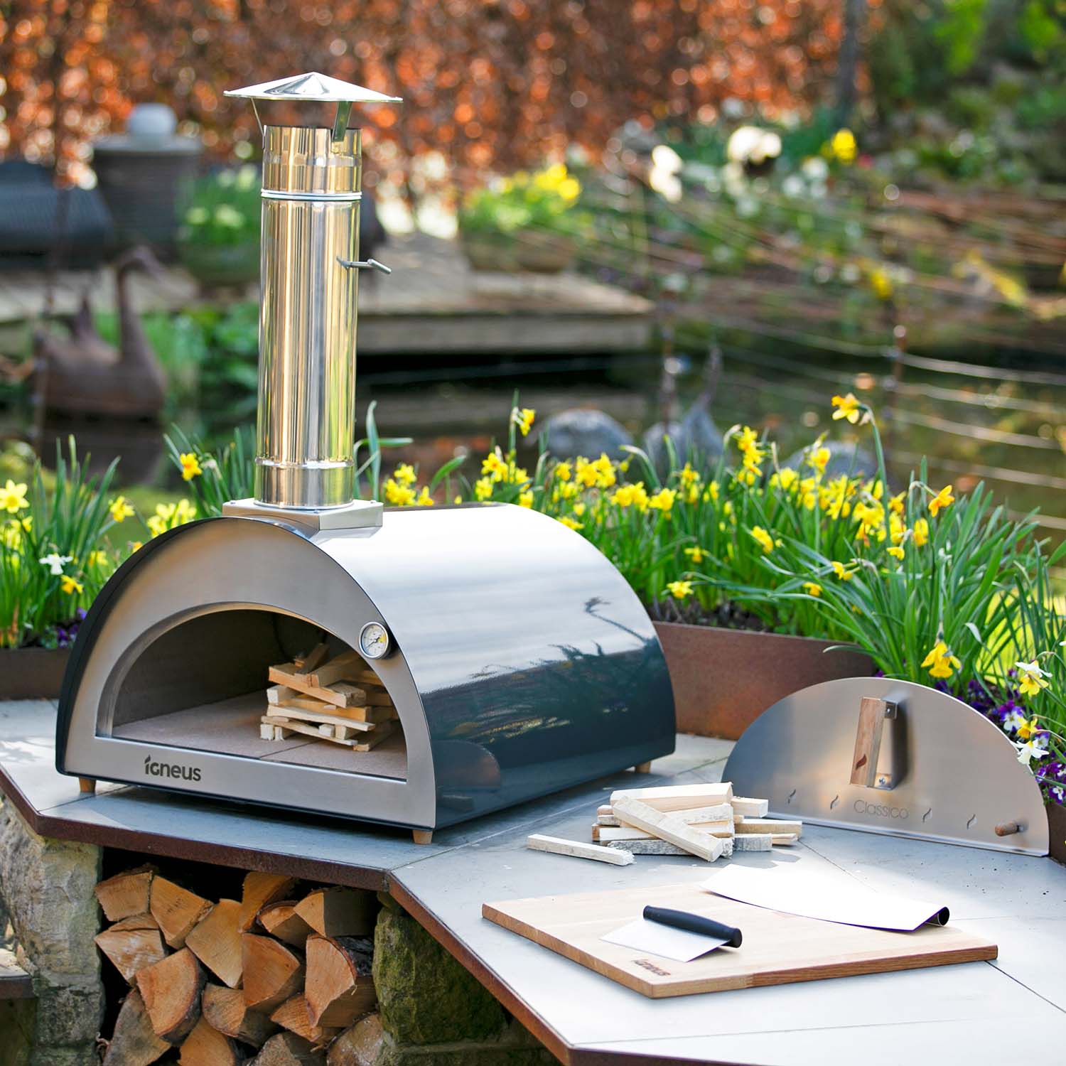 An Igneus Classico Pizza Oven Ultimate Bundle glistens on a stone surface amidst blooming daffodils. Adjacent to the oven, there's a pizza peel and a wooden board. Beneath the oven lies stacked firewood, and atop it extends a tall chimney, ideal for those passionate about outdoor cooking.