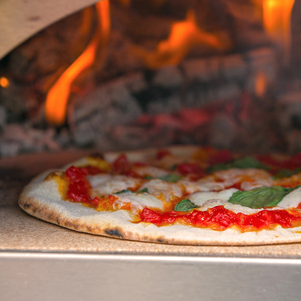 A wood-fired pizza cooked in the Igneus Minimo Portable Pizza Oven (Inc. Peel & Rake) features melted cheese, tomato sauce, and fresh basil leaves. Flames flicker in the background of this outdoor cooking experience, creating a cozy and rustic ambiance.