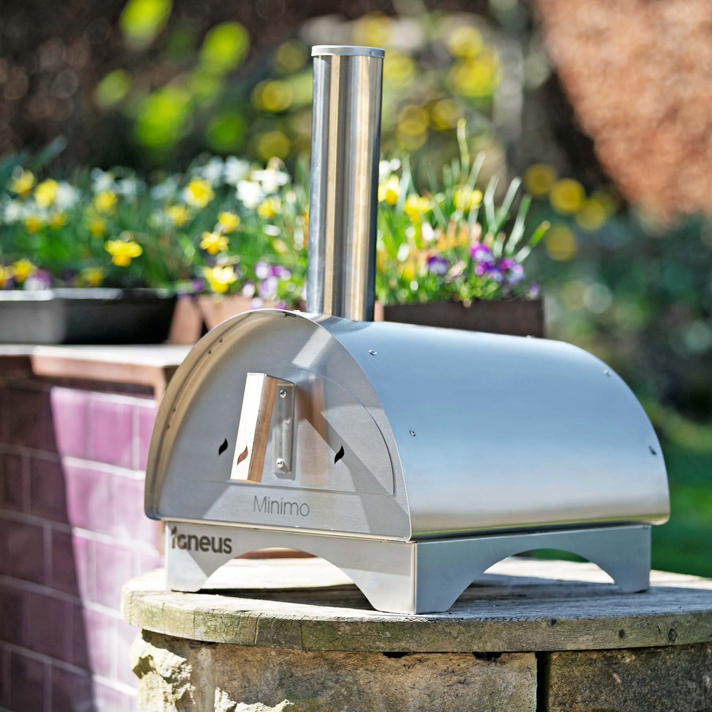 The Igneus Minimo Portable Pizza Oven (Inc. Peel & Rake) stands proudly on a stone surface with its metallic design. A chimney reaches upward as vibrant flowers and lush greenery create the perfect garden setting for outdoor cooking adventures.