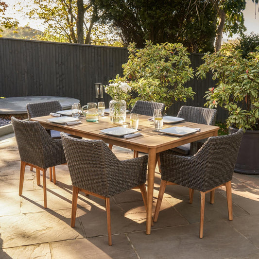 A sunlit patio showcases the Larissa Dining Set Kubu Grey K/D from Pacific Lifestyle, featuring an Acacia wood table accompanied by six faux rattan weave chairs. The table is elegantly arranged with plates, glasses, and a floral centerpiece, all set against a backdrop of lush greenery and a dark fence.