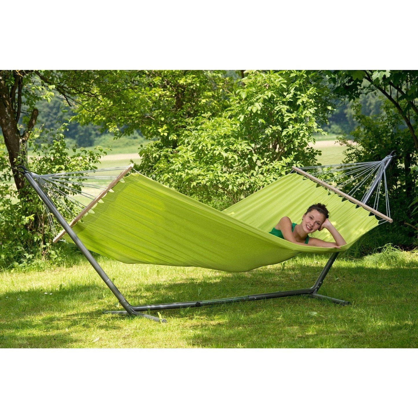 Relaxing on the Amazonas Miami Kiwi Hammock, a person softly sways between its durable spreader bars in a vibrant garden. Resting among trees and grass on its sturdy metal stand, this single hammock offers a peaceful sanctuary of tranquility.