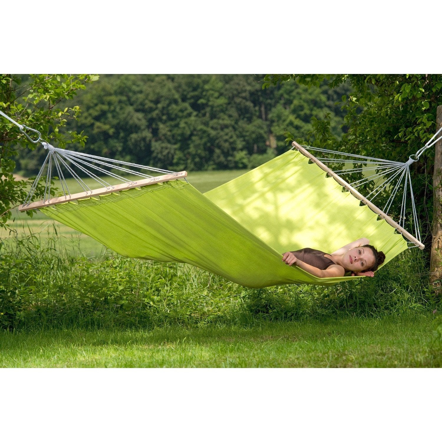 A person relaxes on an Amazonas Miami Kiwi Hammock, securely tied between trees in a grassy area. The green spreader bar enhances stability among lush foliage and open fields under the clear sky.